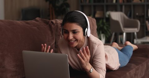 Indian woman lying on sofa look at laptop screen make video call start talking having personal conversation with friend or family living abroad. Female wear headphones communicates by video conference