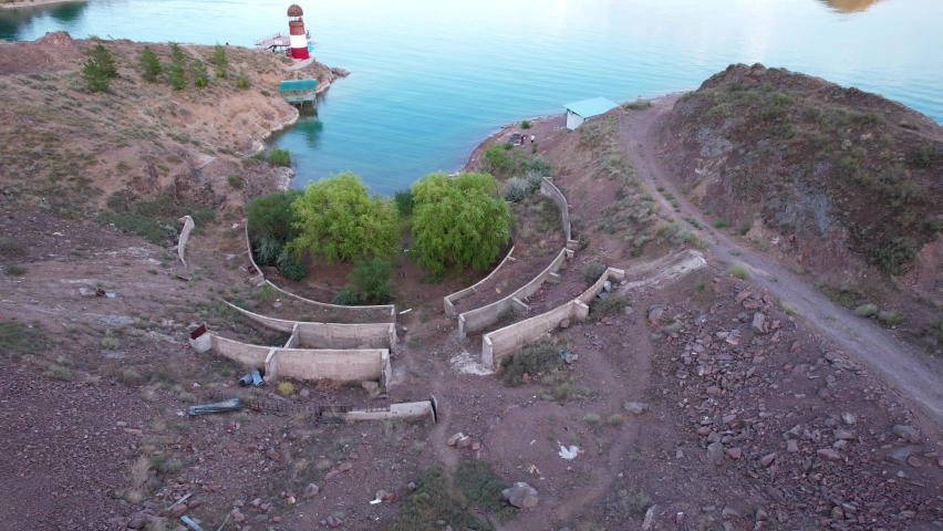 White and red lighthouse on the shore of the lake. The green water of the reservoir reflects the sky and clouds. Rocky beach. In some places, trees and bushes grow. Shtul. Kapchagai, Kazakhstan.