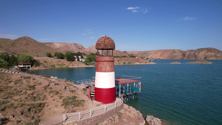 White and red lighthouse on the shore of the lake. The green water of the reservoir reflects the sky and clouds. Rocky beach. In some places, trees and bushes grow. Shtul. Kapchagai, Kazakhstan.