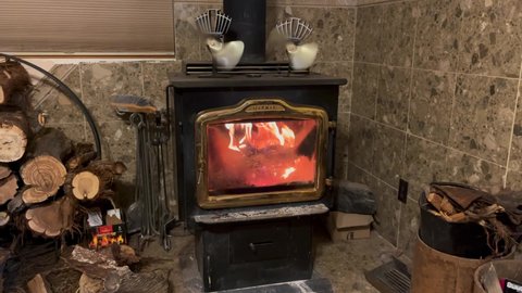 Goshen , Utah , United States - 01 04 2021: A wood burning stove with firewood stacked next to it burns strong with two kinetic fans sitting on top of the stove