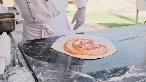 Pizzaiolo putting sauce on dough pizza on the table. Italian food in outdoor restaurant kitchen. Slow motion. Closeup of hands spreading tomato red sauce 