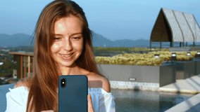 Young Smiling Woman with Smartphone Typing Message or Make Video Call on Rooftop of Hotel near Swimming Pool. Slow Motion of Happy Female Tourist Using Mobile Phone on Sunset