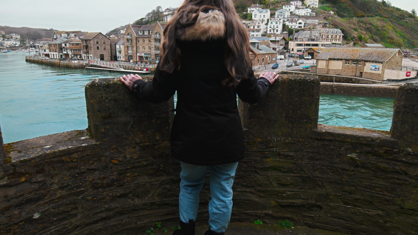 A tourist girl enjoying watching a beautiful location with houses by blue river.  A harbour with boats in old British town Looe with a dream vacation look in Cornwall, England in 4K.