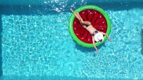 Aerial view of a beautiful woman in bikini taking a sunbath on a watermelon shaped float over blue, sparkling pool water on a hot summer day