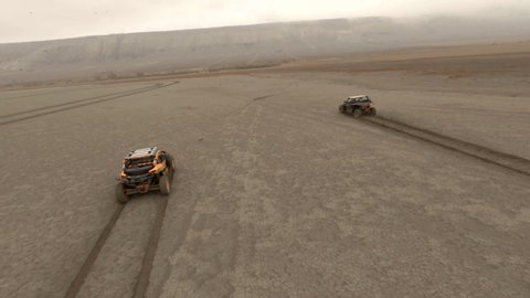 Buggy suv automobiles drive along dried salt lake leaving traces on yellow sand under grey cloudy sky first person chase drone view
