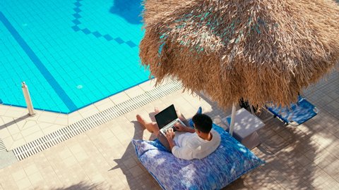 Top View of Man Working on Laptop under Umbrella near Pool in Hotel or Vacation Resort in Tropical Country. Home Office, Male Web Shopping or Freelance Job Concept. 4K Extreme Wide Hand held Shot