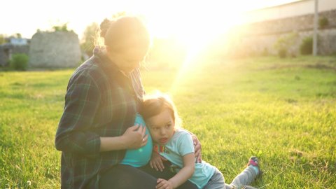 summer, nature, happy family, pregnancy - young pregnant woman mother with two small toddler children sit on grass in park at sunset. Kids stroking mom belly kiss have fun, laugh spend time together
