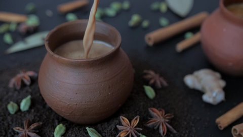 Tasty homemade boiling hot tea poured in traditional clay cups - Indian culture. Famous spices Elaichi, Tej Patta, Dalchini, and cloves kept together against a blurred background - tea-making ingre...