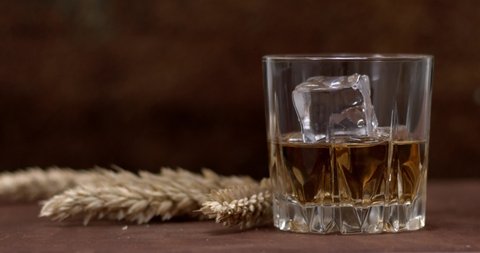 Natural whiskey in a glass with ice cubes on a dark background on a wooden table, near a glass of whiskey, wheat falling. 4K.