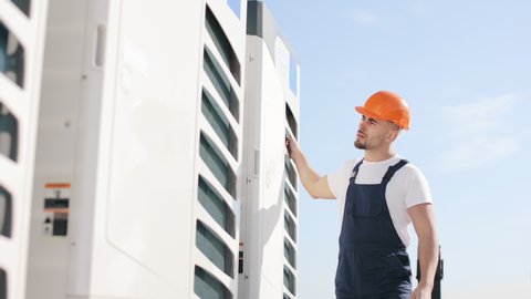 A young engineer is goes and checking the air conditioning system. He is maintaining the air conditioning system. He's on the roof of a business center. He is wearing work clothes and a hard hat. Nice
