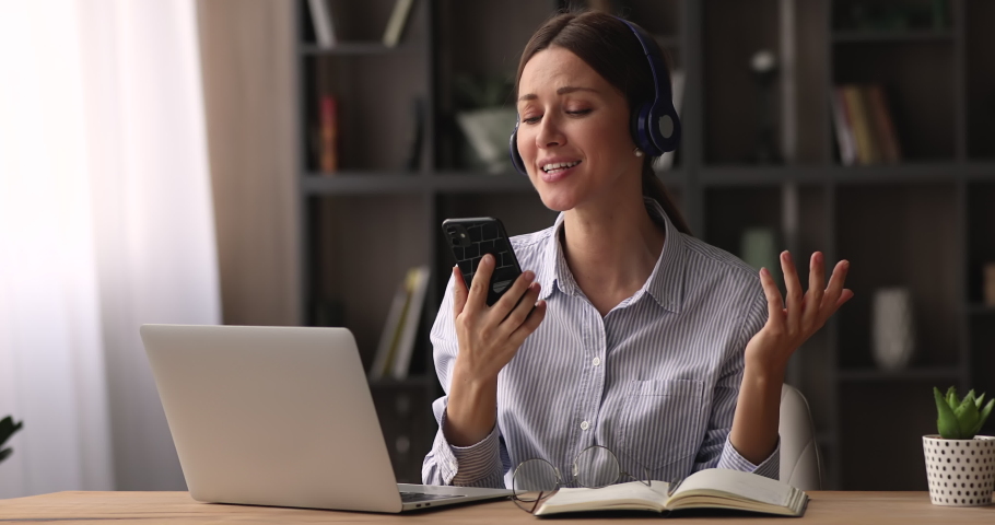 Young woman sit at desk with laptop holding smartphone listen music through wireless headphones watch video clip sing a song enjoy break at workplace. User of online karaoke mobile application concept Royalty-Free Stock Footage #1074180566