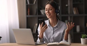 Young woman sit at desk with laptop holding smartphone listen music through wireless headphones watch video clip sing a song enjoy break at workplace. User of online karaoke mobile application concept