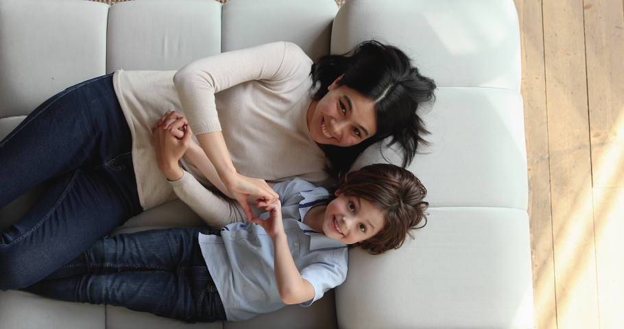 Overhead view happy young Asian mom little Caucasian son lying on couch smile look at camera making heart shape with fingers showing sign of love. Life value, children upbringing and custody concept Royalty-Free Stock Footage #1074180602