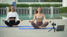 two young fit woman live streaming teach yoga online by swimming pool in sport club outdoors . sporty female practicing online sport class stretching and exercises . workout 