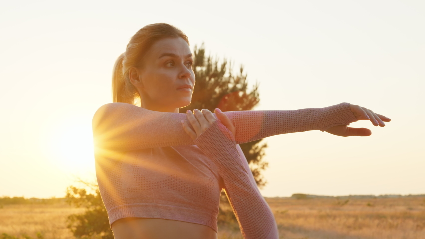Active woman athlete does full body warm-up before training. Sportswoman runner kneads her legs and arms at dawn in summer against background of blue sky on nature of sea. Lifestyle. Sport. Travel