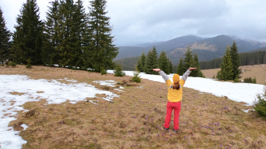 Young girl in yellow hat spins with raised hands in spring mountains and enjoys the beauty of nature landscape with crocuses flowers