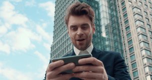 Enthusiastic Man Office Worker Playing Racing Game using Mobile Applications. Attractive Young Employee Rejoices. Wears Suit standing outdoor. Uses Gadget for Video Games. Break time. Winner. Apps.