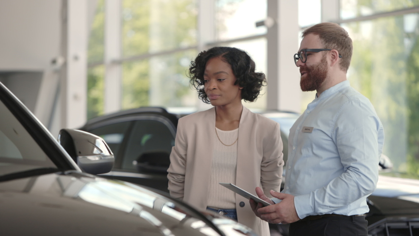 Bearded male dealer using digital tablet while presenting to charming african woman new model of luxury car. Happy female customer choosing new vehicle to buy at showroom. Royalty-Free Stock Footage #1074202934