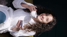 Young curly haired girl drinking a tea at home while is writting a message or watching news on phone after having a bath. The sun shines on her face.