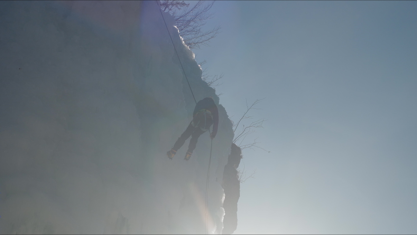 Handheld shot of a female silhouette, ice climber going down an ice waterfall, using a safety top rope, back view