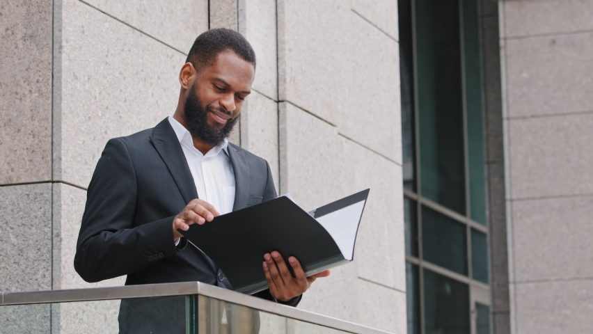 Serious focused African American businessman analyst standing on balcony holding sheets check documents. Mixed race young adult man reading contract papers contract, doing research, prepare report Royalty-Free Stock Footage #1074213614