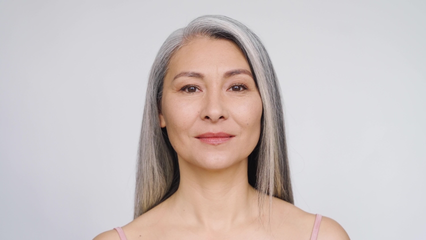 Middle aged mature asian woman, senior older 50 year lady looking at camera smiling touching face applying antiaging uv protection whitening skin care cream, closeup face portrait isolated on white. | Shutterstock HD Video #1074214259