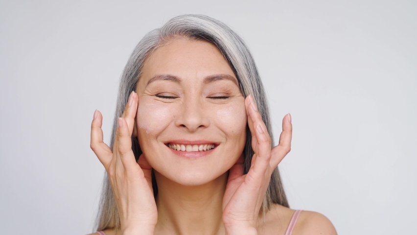 Middle aged mature asian woman, senior older 50 year lady looking at camera smiling touching face applying antiaging uv protection whitening skin care cream, closeup face portrait isolated on white. Royalty-Free Stock Footage #1074214259
