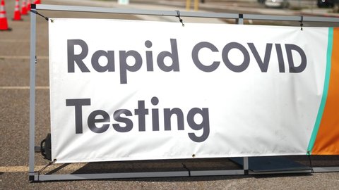 Rapid Covid 19 Testing site sign posted outside on sunny summer day in empty parking lot with orange cones setup, and cars driving by in background. In 4k slow motion.