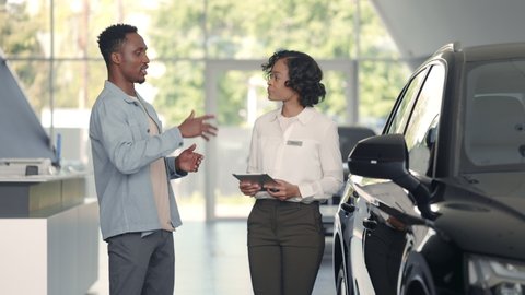 Female manager with digital tablet in hands working with male customer at auto salon. African woman in formal clothes presenting car to young man.