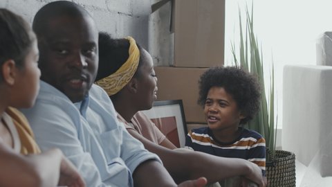 Side view shot PAN of African-American parents and kids sitting on floor in their new home and chatting Their belongings are in cardboard boxes
