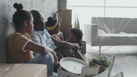 Side view shot of happy African-American parents and children sitting together on floor of their new apartment and chatting Their belongings are packed in boxes