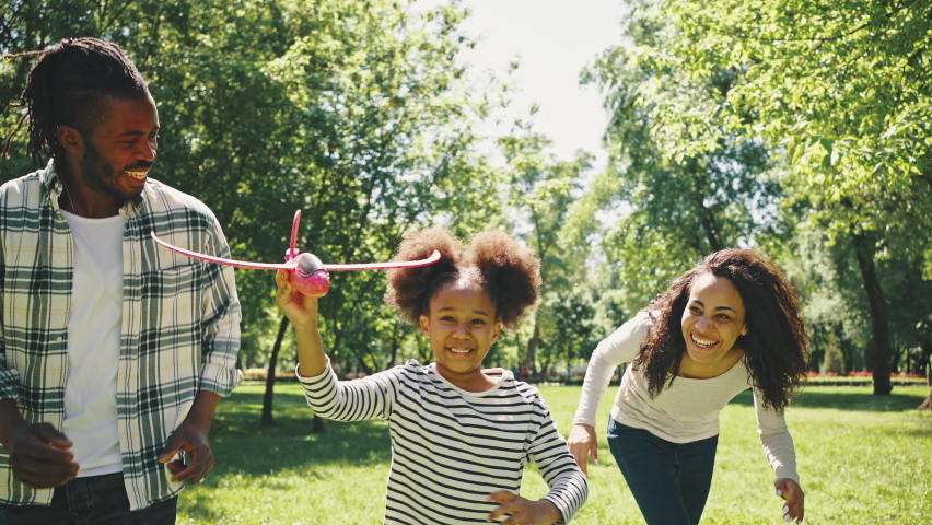 Cute black girl and her parents are playing in the park with a red plane. African american family have fun during a weekend in nature. Royalty-Free Stock Footage #1074250376