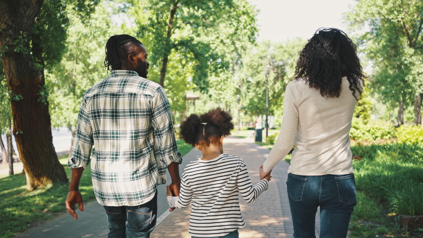 Back view of smiling dark-skinned man and woman walk in the park holding the hands of their little daughter. Being together, family weekend, picnic in the park.
