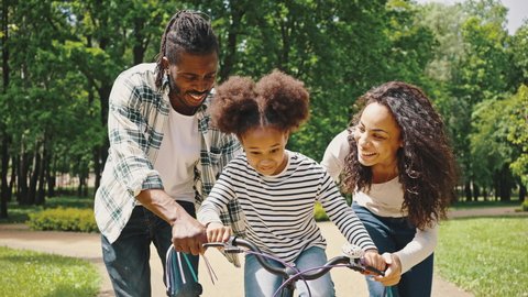 Happy family with excited daughter learning to ride a bike for the first time. African American parents teaching their little girl to driving bike in park.