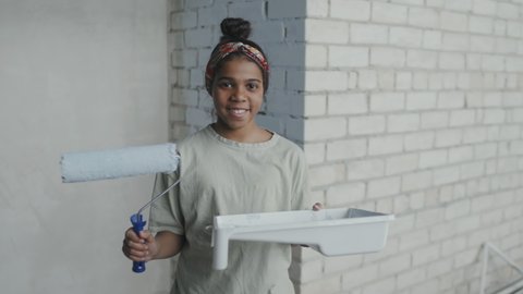 PAN slowmo portrait shot of happy African-American girl holding paint roller and tray and posing for camera inside apartment with brick wall