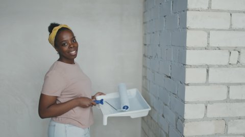 Portrait slowmo shot of happy African-American woman painting brick wall and posing for camera while renovating house