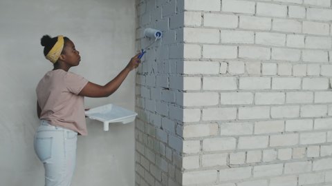 Side view shot of African-American woman painting brick wall while renovating house