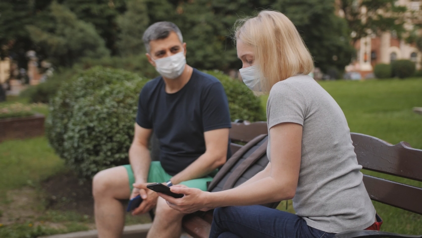 A man and a woman wearing protective medical masks sit on opposite parts of a park bench and communicate.
