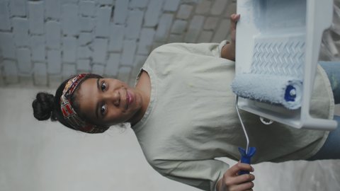 Vertical portrait of happy African-American girl with paint roller and tray posing for camera in apartment with brick wall