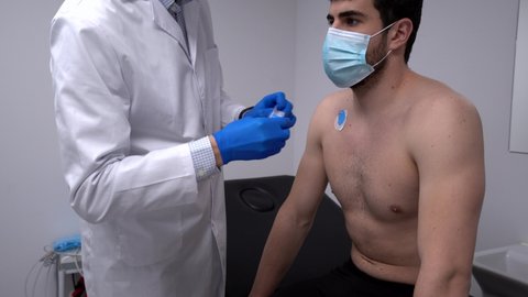 Crop medic applying electrodes on chest of unrecognizable male patient in mask for ECG test