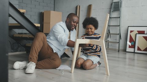 Medium shot of happy African-American father and son smiling and assembling wooden chair in loft apartment