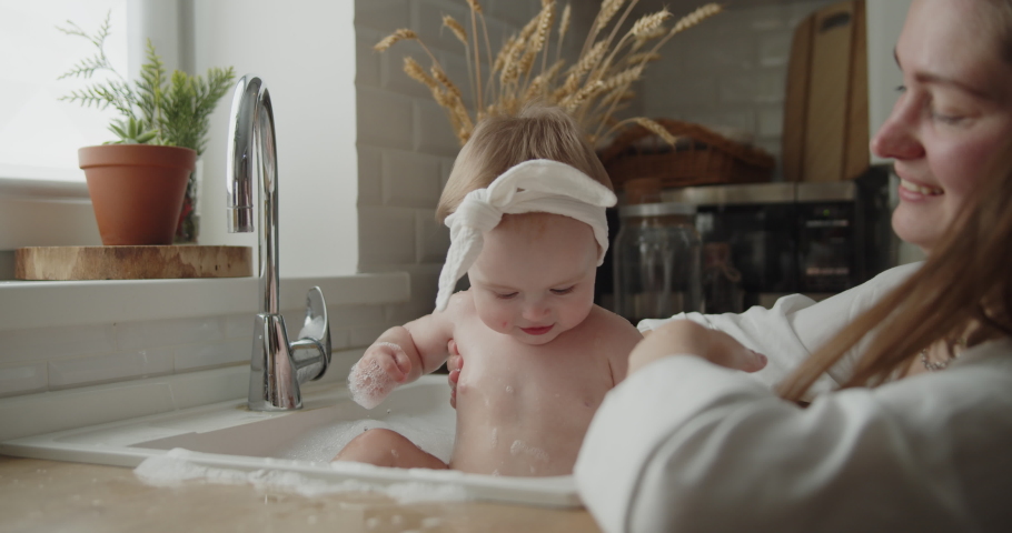 Mother and daughter spend time at home together. Young mom helps her adorable little child to take a bath in a kitchen sink. Adorable girl have fun in the water and foam. Royalty-Free Stock Footage #1074264755
