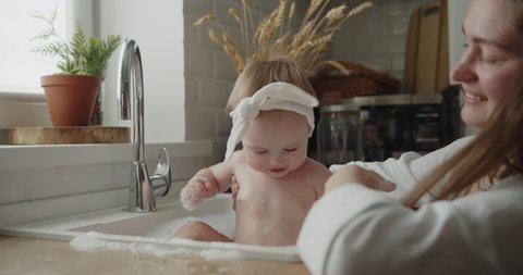 Mother and daughter spend time at home together. Young mom helps her adorable little child to take a bath in a kitchen sink. Adorable girl have fun in the water and foam.