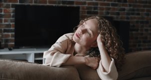 Blonde woman with curly hair leans over a sofa, smiling, resting her chin in her hands. Against the background of a chic living room. High quality 4k footage