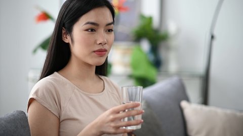 Healthy lifestyles. Satisfied young asian brunette pretty woman sitting at home on sofa in t-shirt, leading a healthy lifestyle, drinking a glass of pure water, smiling. Drink daily norm of water