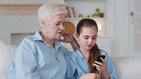 Little cute granddaughter holds smartphone in hands teaches old grandmother to use mobile application, play games and shows photos. Elderly aged babysitter helps girl with education video on cellphone