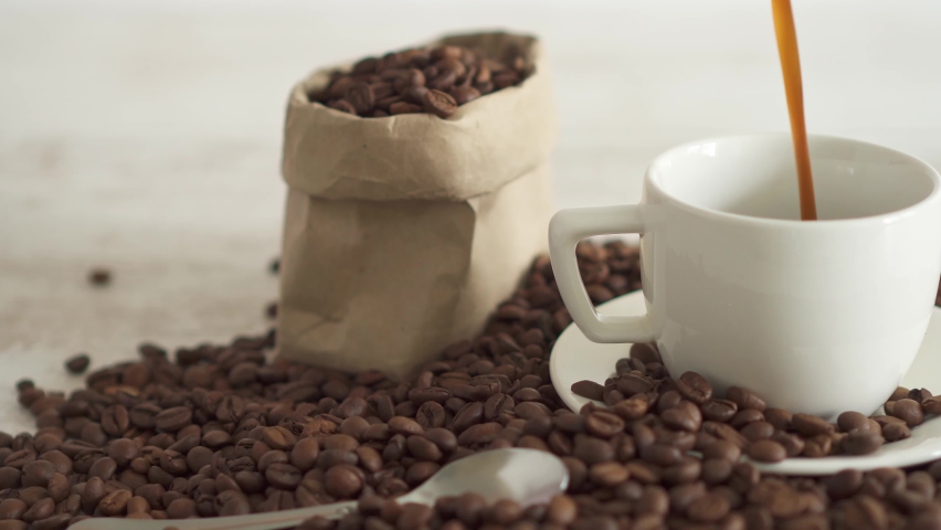 Black hot coffee is poured into the coffee cup. Hot espresso fills a white cup with a huge pile of roasted coffee beans in the background. Storing coffee beans in a paper bag. Falling boiling water  Royalty-Free Stock Footage #1074301370