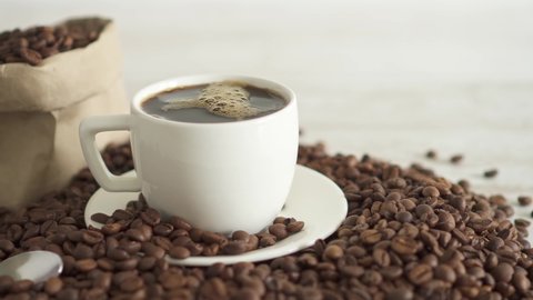 Black hot coffee is poured into the coffee cup. Hot espresso fills a white cup with a huge pile of roasted coffee beans in the background. Storing coffee beans in a paper bag. Falling boiling water 