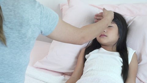 Asian mother using hand checking temperature of sickness little daughter on the bed in bedroom. Mom caring illness child girl kid at home. Family relationship and medical health insurance concept