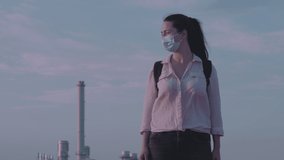 Brunette long hair girl in pink shirt, anti virus mask in pandemic time stand still alone watch around with urban construction factory background. Atmospheric low key video.
